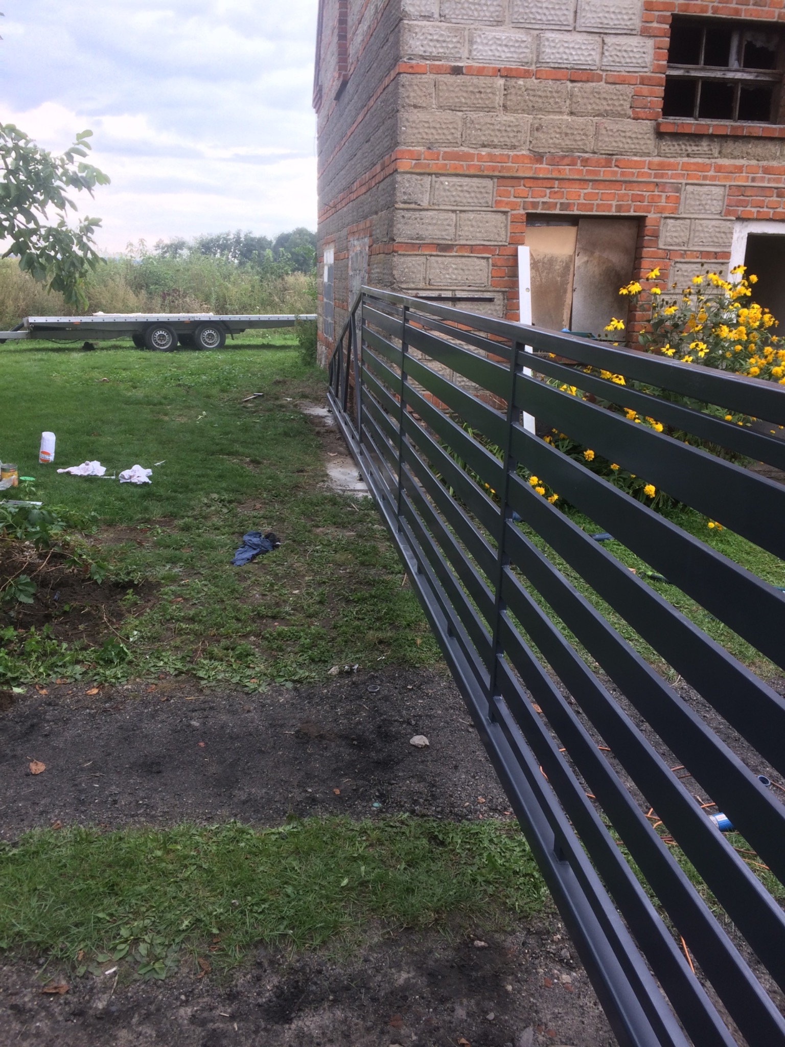 Nowoczesna, pozioma brama wjazdowa z metalowych profili, świeżo pomalowana na ciemnoszary kolor, ustawiona przy ceglanym budynku w trakcie budowy, z widocznym przyczepą i polnymi kwiatami w tle.