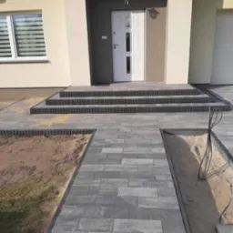 Nowoczesny podjazd z szarej kostki brukowej prowadzący do schodów wejściowych domu z białymi drzwiami.