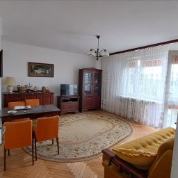 PROPERTON SPÓŁKA Z OGRANICZONĄ ODPOWIEDZIALNOŚCIĄ - Widok na umeblowany salon z drewnianą podłogą, owalnym dywanem, stołem z pomarańczowymi krzesłami, komodą, witryną i telewizorem, widziany zza otwartych drzwi.