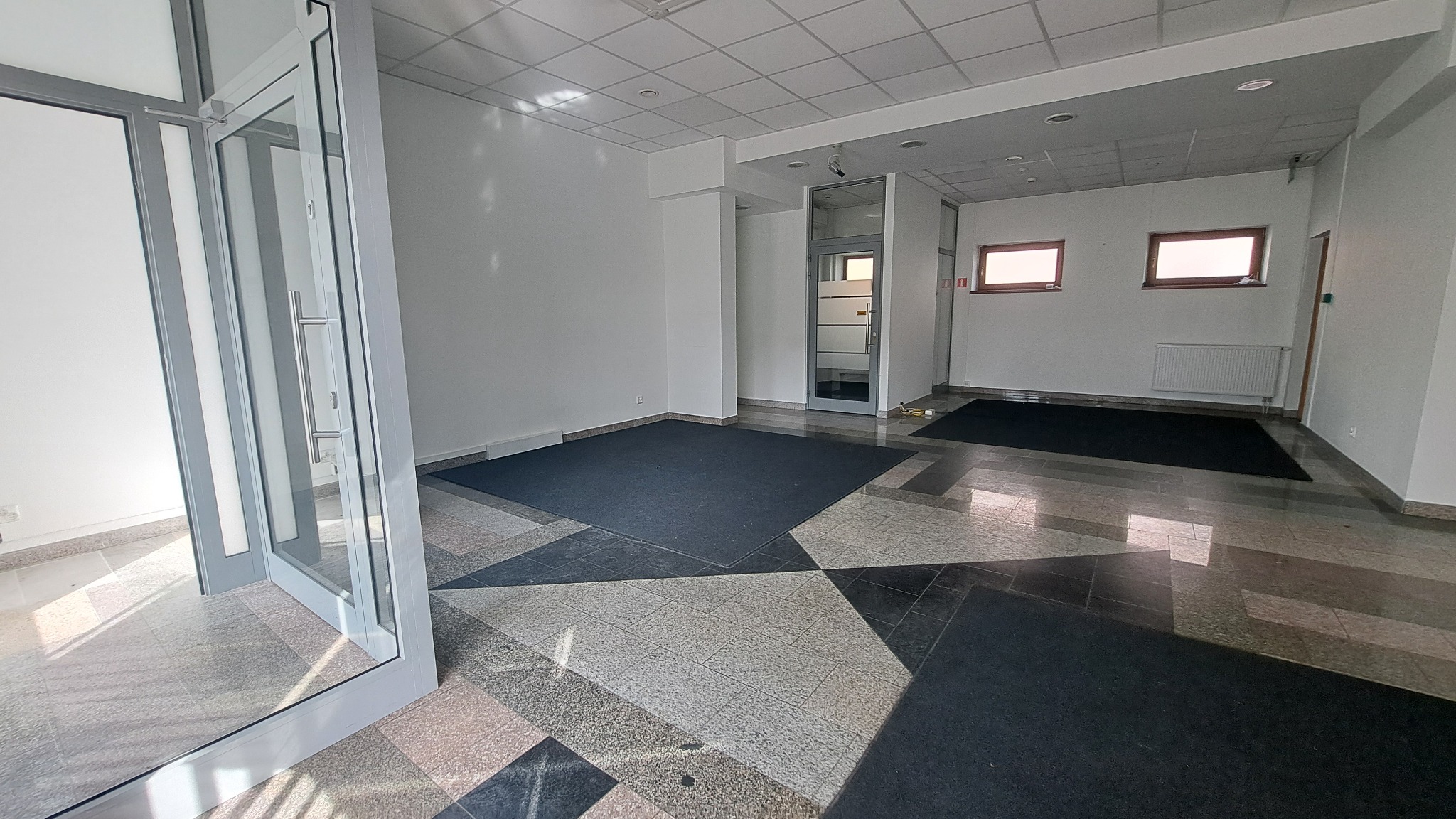 Puste, jasne pomieszczenie biurowe z otwartymi szklanymi drzwiami wejściowymi, ciemnymi wycieraczkami i mozaikową podłogą.
