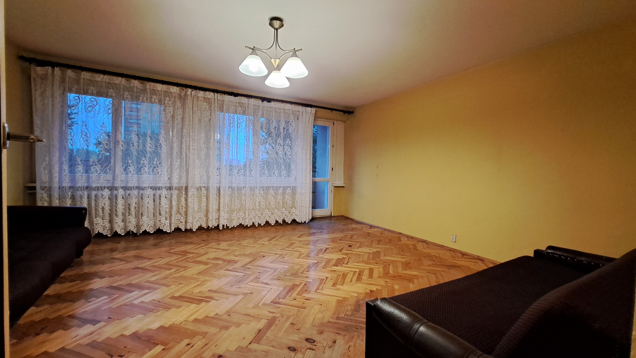 Pusta przestrzeń w mieszkaniu z parkietem w jodełkę, zasłoniętym oknem i żółtymi ścianami, widoczny fragment ciemnej sofy.