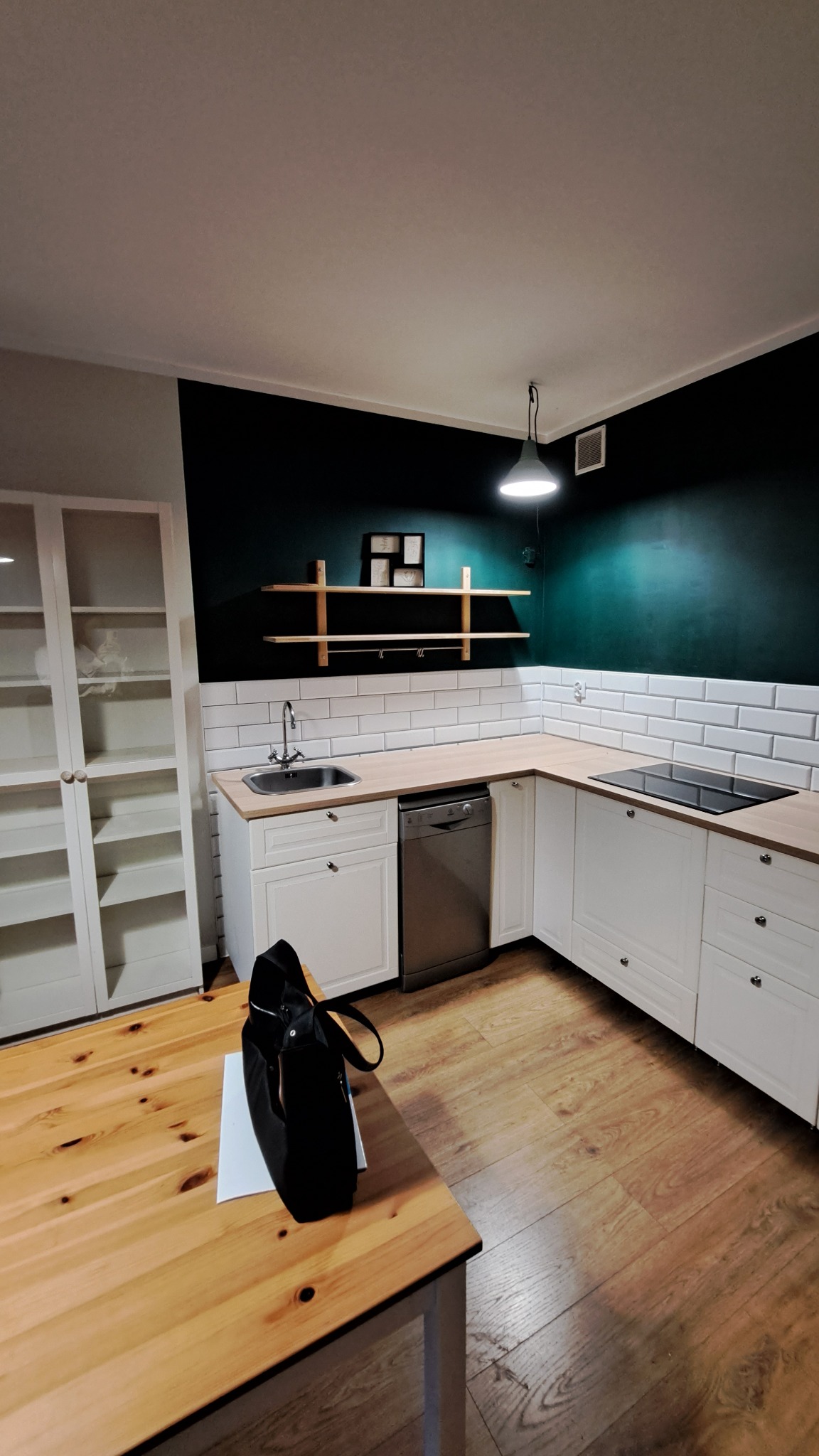 Nowoczesna kuchnia z białymi szafkami, drewnianym blatem i ciemnozieloną ścianą, widziana z perspektywy stołu z czarną torbą.