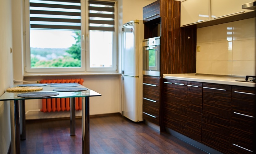 Jasna kuchnia z ciemnymi drewnianymi szafkami, szklanym stołem z podkładkami, białym lodówką i pomarańczowym grzejnikiem pod oknem z żaluzjami.