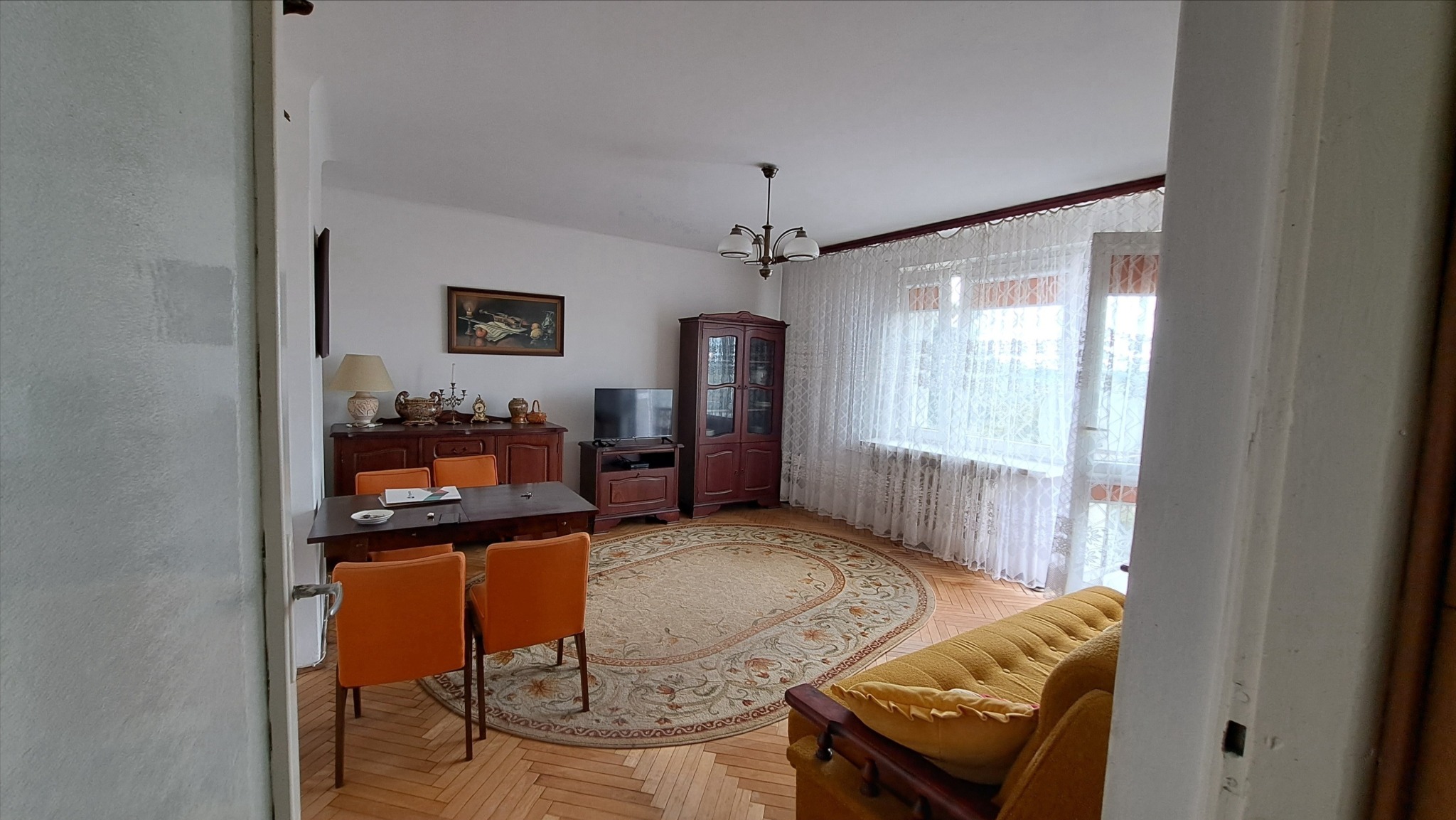 Widok na umeblowany salon z drewnianą podłogą, owalnym dywanem, stołem z pomarańczowymi krzesłami, komodą, witryną i telewizorem, widziany zza otwartych drzwi.