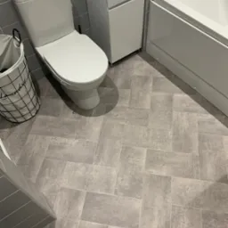 Nowoczesna łazienka w odcieniach szarości z płytkami ułożonymi w jodełkę, biała toaleta z opakowaniem chusteczek na spłuczce, biała szafka i wanna.