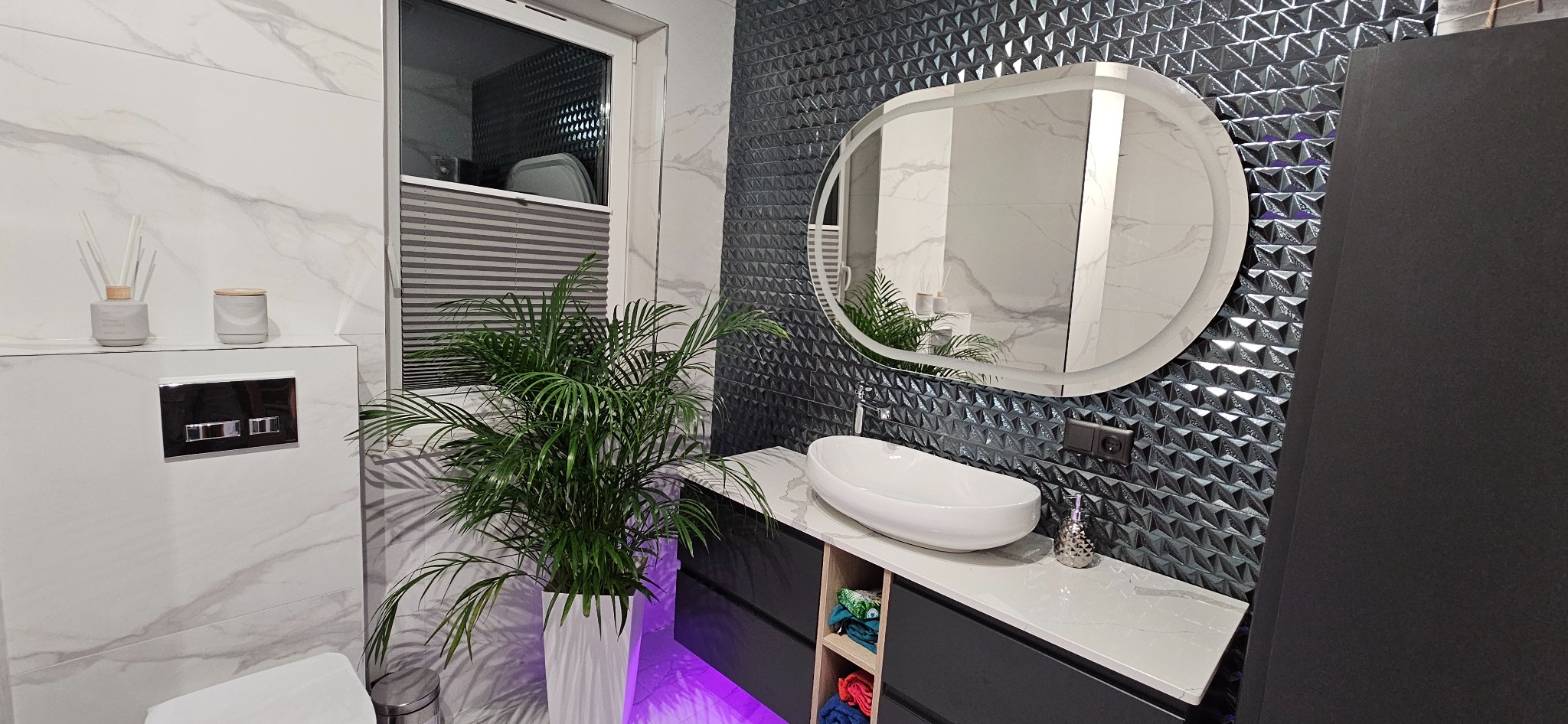 Nowoczesna łazienka z umywalką nablatową, podświetlanym lustrem owalnym i ścianą wyłożoną trójwymiarowymi płytkami w odcieniach szarości, oświetlona fioletowym światłem od dołu.