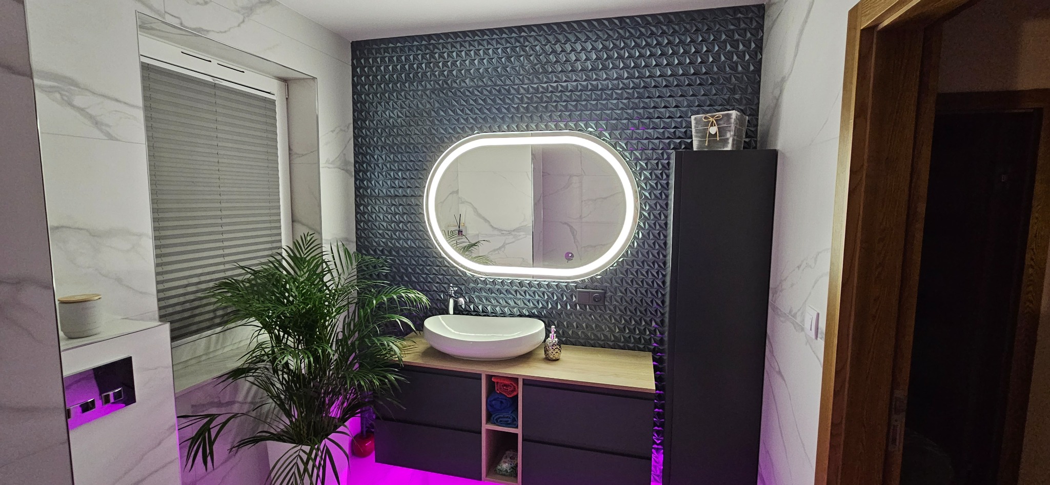 Nowoczesna łazienka z podświetlanym lustrem owalnym, umywalką nablatową, szafką z drewnianym blatem, szarą ścianą z trójwymiarową fakturą, marmurowymi płytkami i rośliną doniczkową.