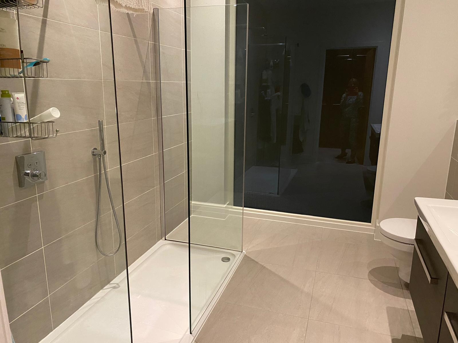 Nowoczesna łazienka z kabiną prysznicową typu walk-in z czarnym szkłem, szare płytki na ścianach i podłodze, chromowana armatura, widoczne kosmetyki w koszyczku i odbicie osoby w lustrze.