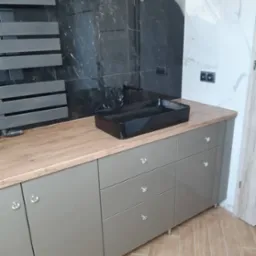 Szara szafka łazienkowa z drewnianym blatem i czarną umywalką na tle czarno-białych płytek ceramicznych i grzejnika drabinkowego.