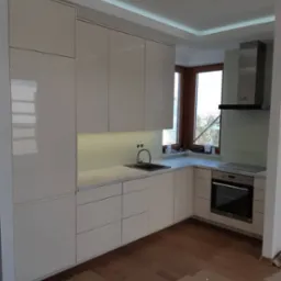 Jasna, nowoczesna kuchnia z białymi, lakierowanymi frontami, szarym blatem, wbudowanym oświetleniem podszafkowym i okapem ze stali nierdzewnej, widok na narożne okno z drewnianą ramą.