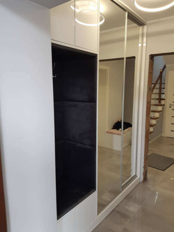Biała szafa wnękowa z lustrzanymi drzwiami przesuwnymi i otwartą wnęką z ciemnoszarą tapicerowaną ścianką, widok na korytarz z schodami.