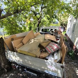 Oleksandra Hoiarchuk Złote Drzewo - Ciężarówka wypełniona po brzegi różnymi odpadami, w tle zieleń drzew i fragment budynku. Widoczne meble, kartony i inne przedmioty przeznaczone do utylizacji.