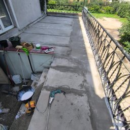Oleksandra Hoiarchuk Złote Drzewo - Betonowy balkon po wylewce, widoczne narzędzia: mieszadło elektryczne, wiadro z zaprawą. W tle barierka, zieleń i zabudowania. Prace wykończeniowe na balkonie.