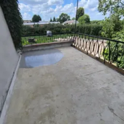 Balkon w trakcie renowacji: częściowo wylana, szara masa wyrównująca, czarna, ozdobna balustrada, widok na zieleń i błękitne niebo z chmurami.