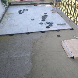 Oleksandra Hoiarchuk Złote Drzewo - Układanie płytek na tarasie z wykorzystaniem podkładek dystansowych. Część płytek ułożona, widoczny poziomica, paczka z płytkami i fragment osoby w kadrze.
