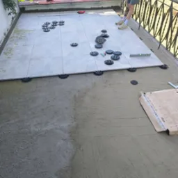 Układanie płytek na tarasie z wykorzystaniem podkładek dystansowych. Część płytek ułożona, widoczny poziomica, paczka z płytkami i fragment osoby w kadrze.