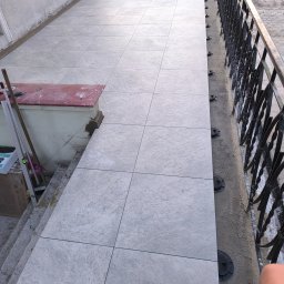 Oleksandra Hoiarchuk Złote Drzewo - Jasnoszare płytki tarasowe ułożone na regulowanych wspornikach, widoczne narzędzia i poziomica. Balustrada z kutego żelaza w tle. Częściowo ukończona realizacja.