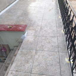 Oleksandra Hoiarchuk Złote Drzewo - Ułożona, jasna kostka brukowa na tarasie z czarną, metalową balustradą. Widoczne fragmenty schodów i ściany budynku. Na kostce żółte plamy po taśmie malarskiej.