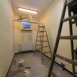 Złote Drzewo Wycinka Drzew Ogrody Budowa - Wnętrze w trakcie malowania: drabiny, puszka farby Śnieżka, narzędzia malarskie na podłodze. Ściany w odcieniach beżu, widoczny grzejnik i okno. Przygotowanie do malowania.
