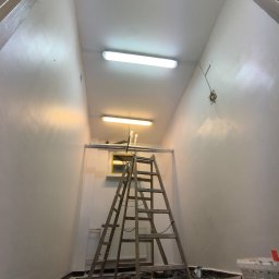 Złote Drzewo Wycinka Drzew Ogrody Budowa - Wnętrze klatki schodowej w trakcie malowania. Widoczna drabina, zabezpieczona podłoga, świeżo pomalowane ściany i sufit. Dwa źródła światła jarzeniowego.