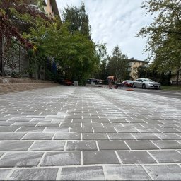 Złote Drzewo Wycinka Drzew Ogrody Budowa - Nowo ułożona szara nawierzchnia z betonowych elementów, z białym piaskiem w spoinach, widok z niskiej perspektywy. W tle pracownik z taczką, samochody i budynki.