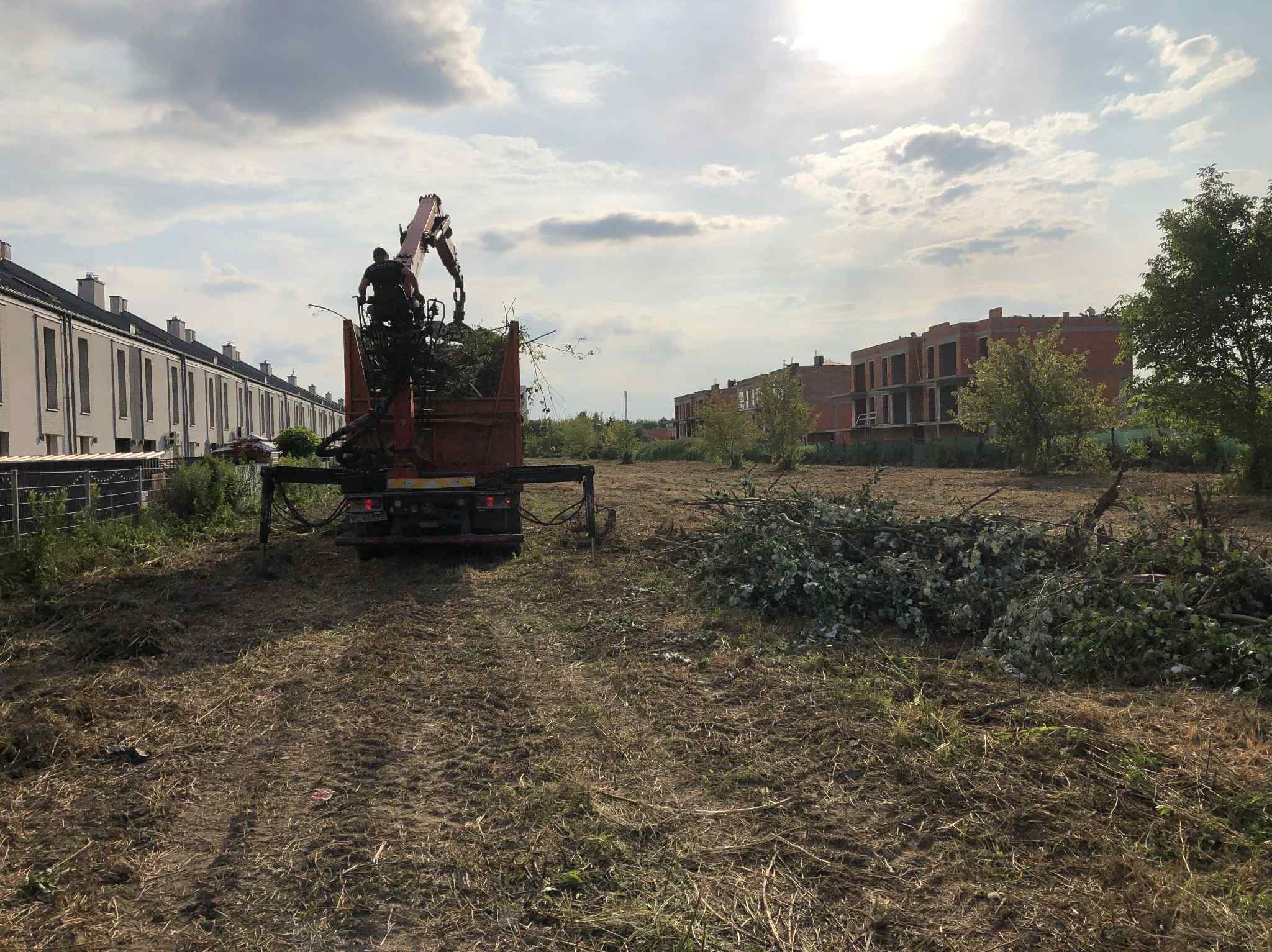 Ciężarówka z dźwigiem załadowana gałęziami na tle zabudowy miejskiej. Porządkowanie terenu po wycince. Słoneczny dzień, lekko zachmurzone niebo.