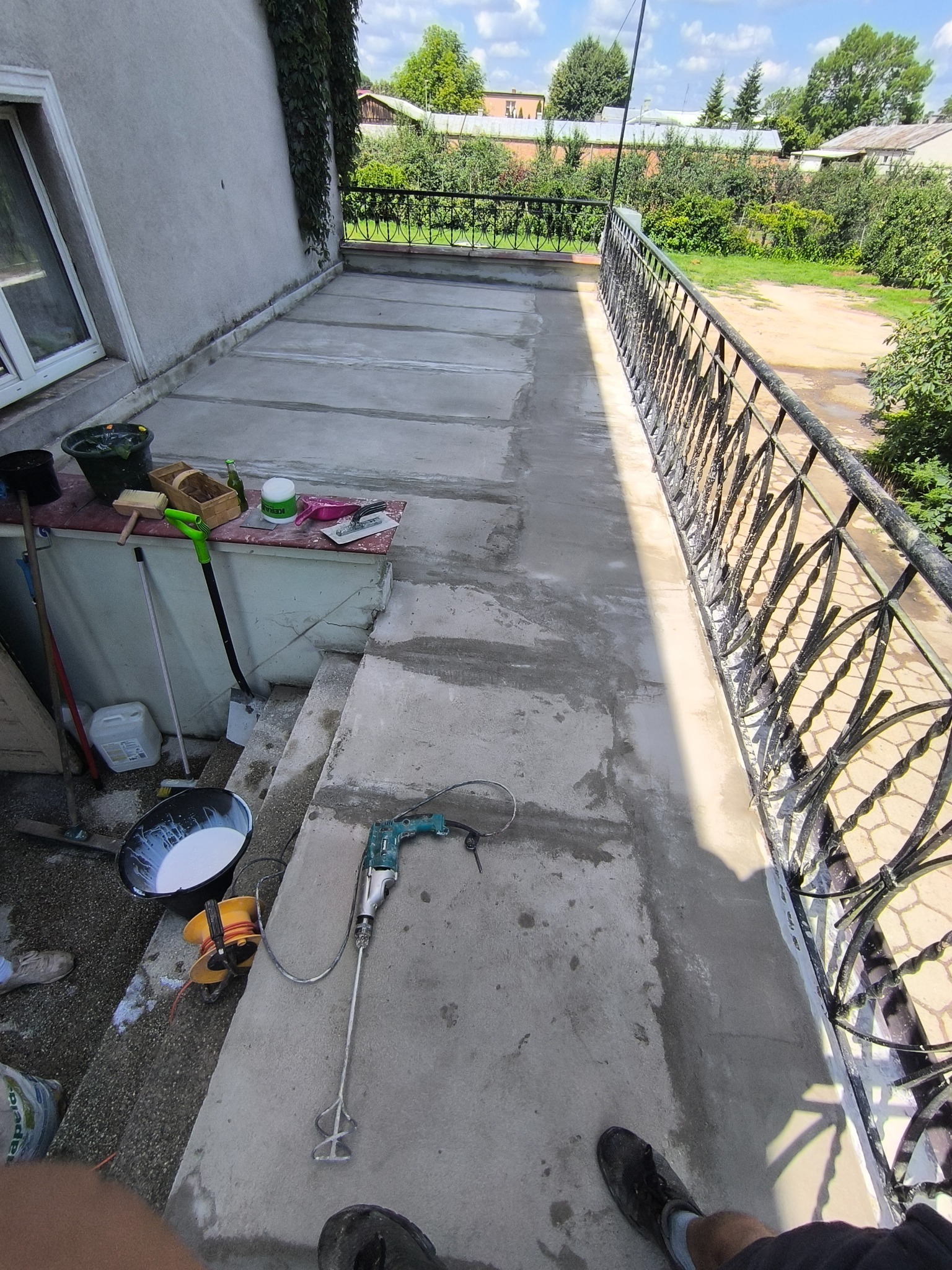 Betonowy balkon po wylewce, widoczne narzędzia: mieszadło elektryczne, wiadro z zaprawą. W tle barierka, zieleń i zabudowania. Prace wykończeniowe na balkonie.