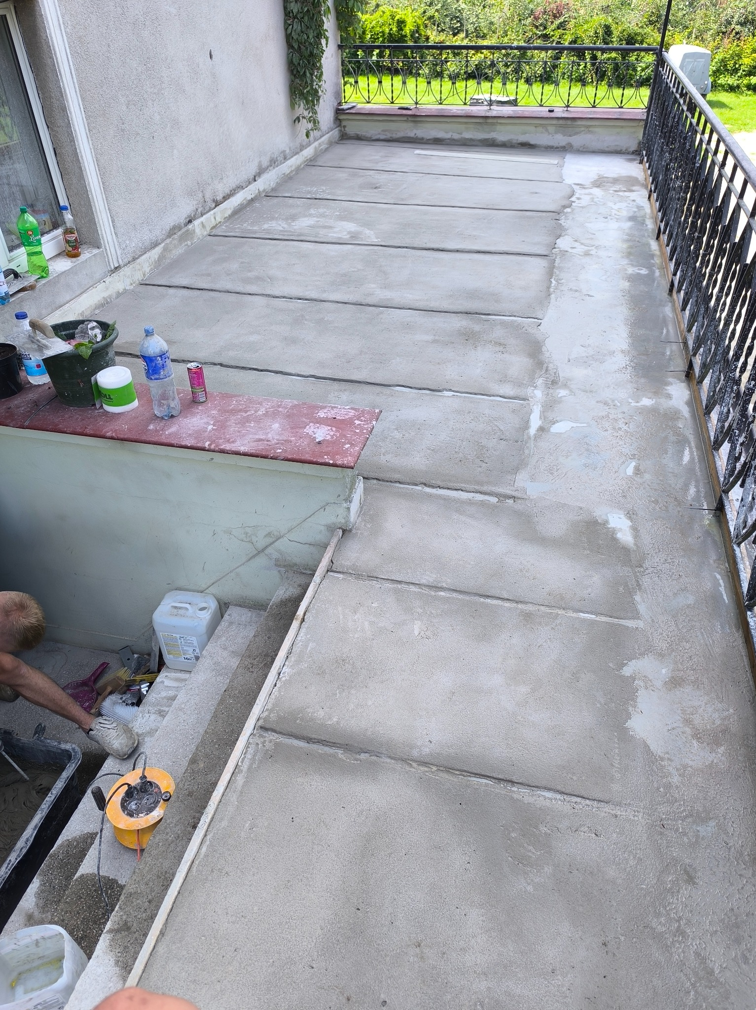 Wyrównywanie powierzchni betonowej na tarasie w Grójcu. Widoczny fragment balustrady i narzędzia pracy. Prace remontowe na zewnątrz budynku.