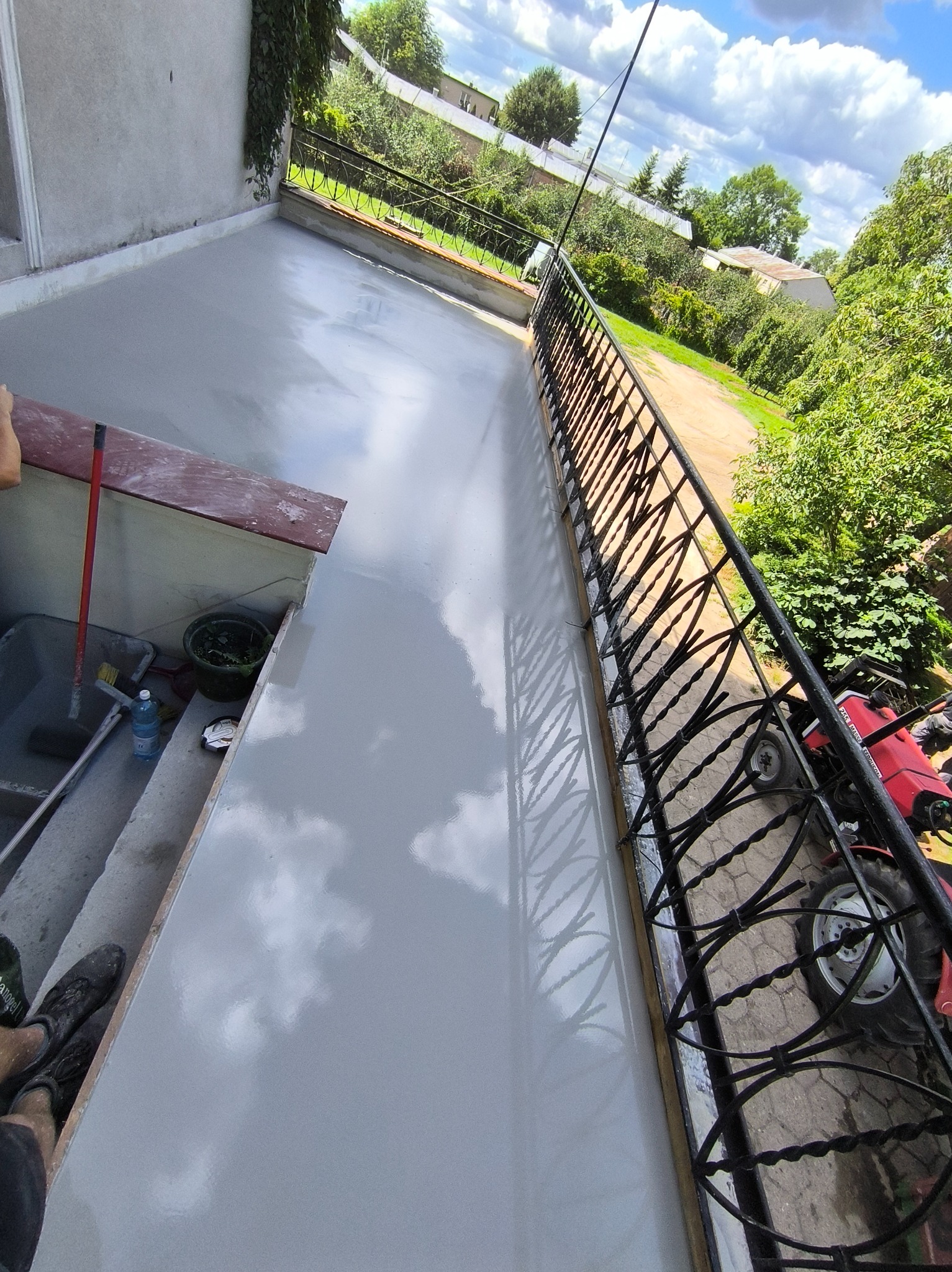 Wylany beton na balkonie z czarną balustradą. Widoczne narzędzia pracy, wiadro, szczotka. W tle zieleń i niebo z chmurami. Praca w toku, świeży beton.