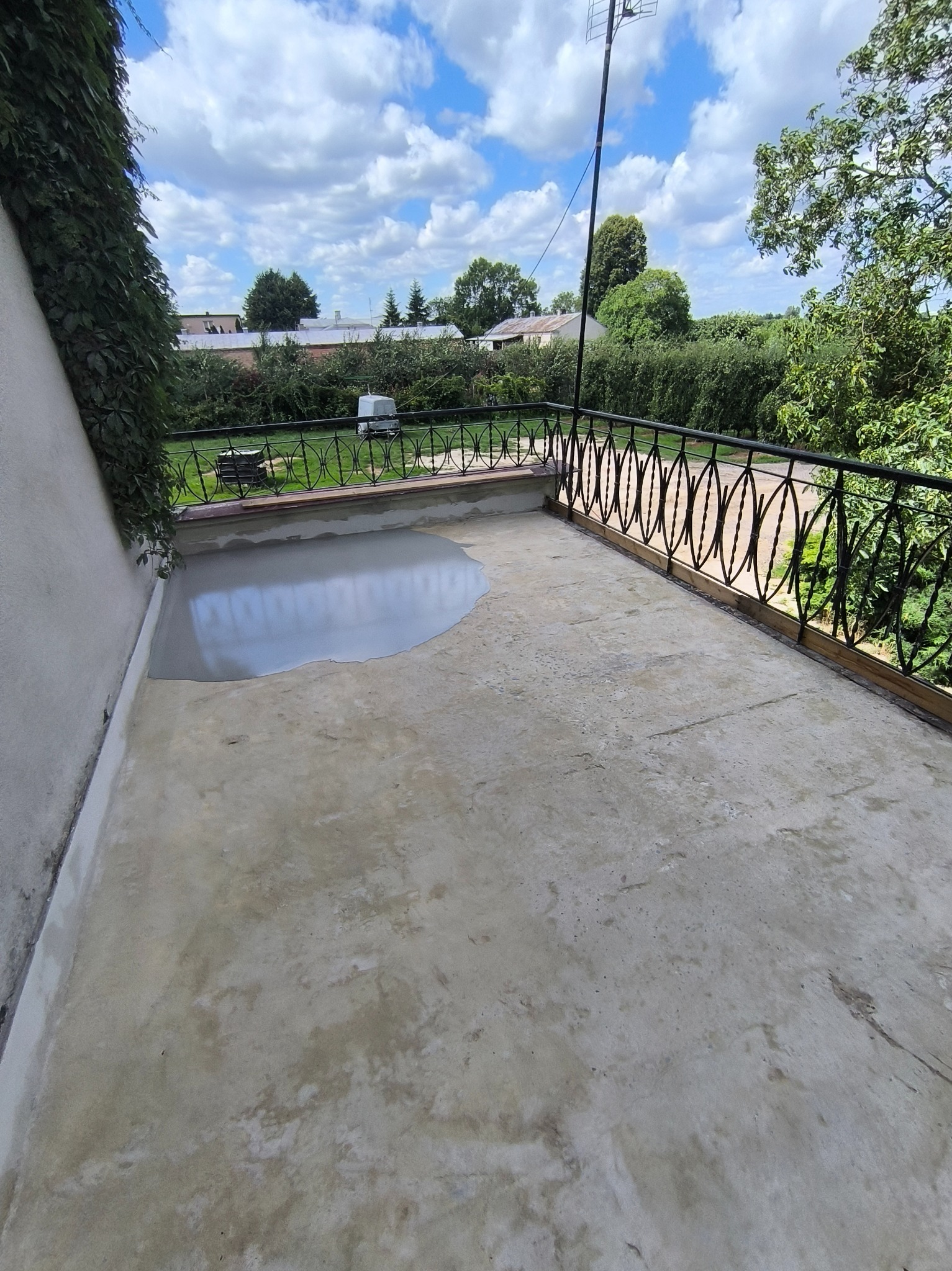 Balkon w trakcie renowacji: częściowo wylana, szara masa wyrównująca, czarna, ozdobna balustrada, widok na zieleń i błękitne niebo z chmurami.