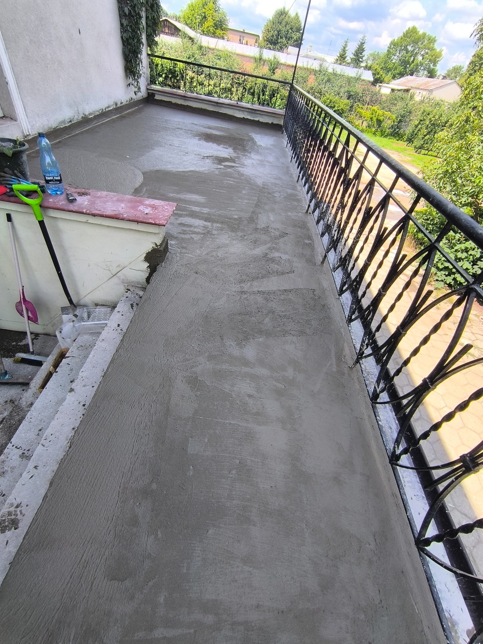 Wylany beton na balkonie z kutą balustradą. Widoczne narzędzia i butelka wody. Świeża wylewka, gotowa do dalszych prac wykończeniowych. Perspektywa z góry.