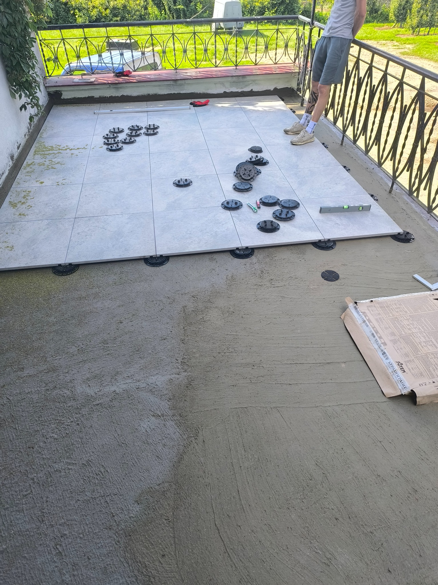 Układanie płytek na tarasie z wykorzystaniem podkładek dystansowych. Część płytek ułożona, widoczny poziomica, paczka z płytkami i fragment osoby w kadrze.