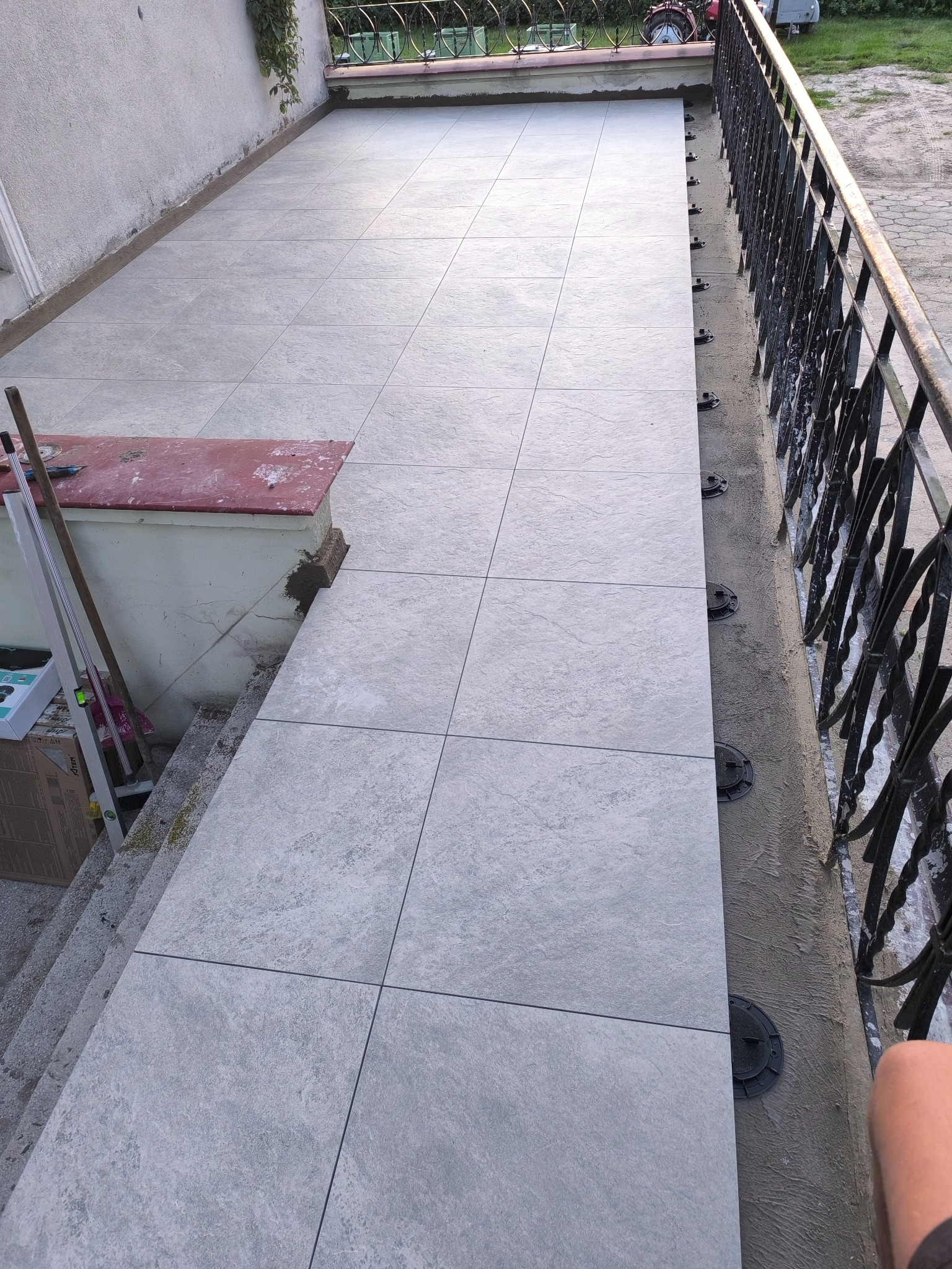 Jasnoszare płytki tarasowe ułożone na regulowanych wspornikach, widoczne narzędzia i poziomica. Balustrada z kutego żelaza w tle. Częściowo ukończona realizacja.