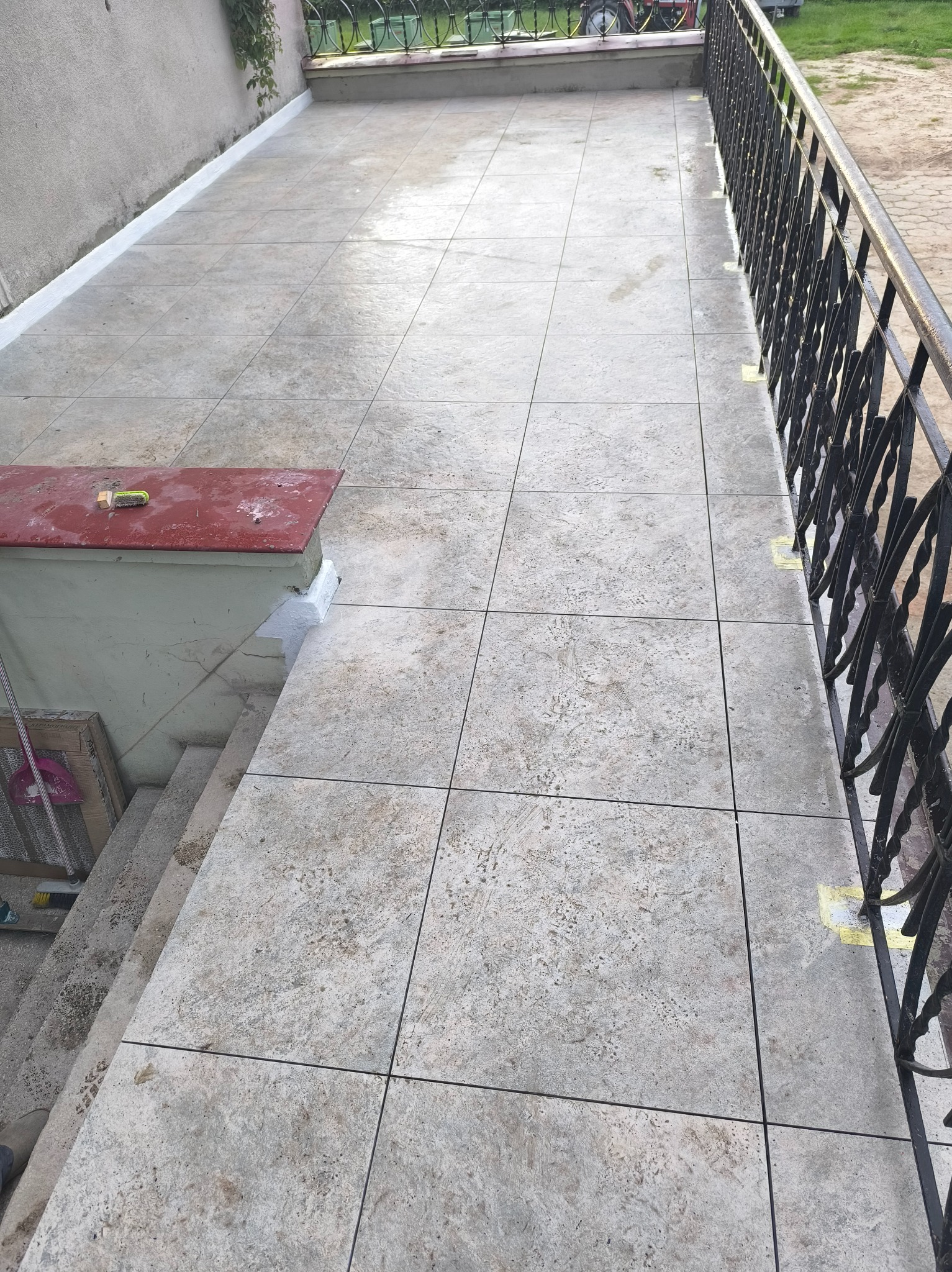 Ułożona, jasna kostka brukowa na tarasie z czarną, metalową balustradą. Widoczne fragmenty schodów i ściany budynku. Na kostce żółte plamy po taśmie malarskiej.