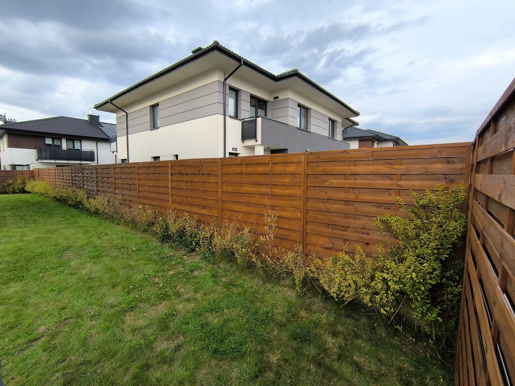 Nowoczesny, drewniany płot panelowy oddzielający posesję, widok z trawnika. Tło stanowi dwukondygnacyjny dom z balkonem i sąsiednie budynki pod pochmurnym niebem.
