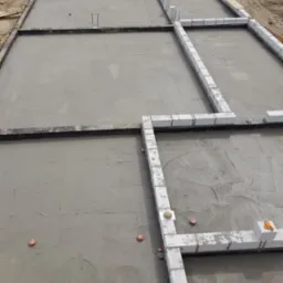 Zalany betonowy fundament z wyznaczonymi strefami pod ściany z bloczków Ytong, widoczne pomarańczowe znaczniki i wystające pręty zbrojeniowe.