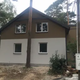 Dwukondygnacyjny dom z ocieploną elewacją w kolorach szarości i brązu, widoczny pień drzewa na pierwszym planie, oraz skrzynka elektryczna na słupie obok budynku.