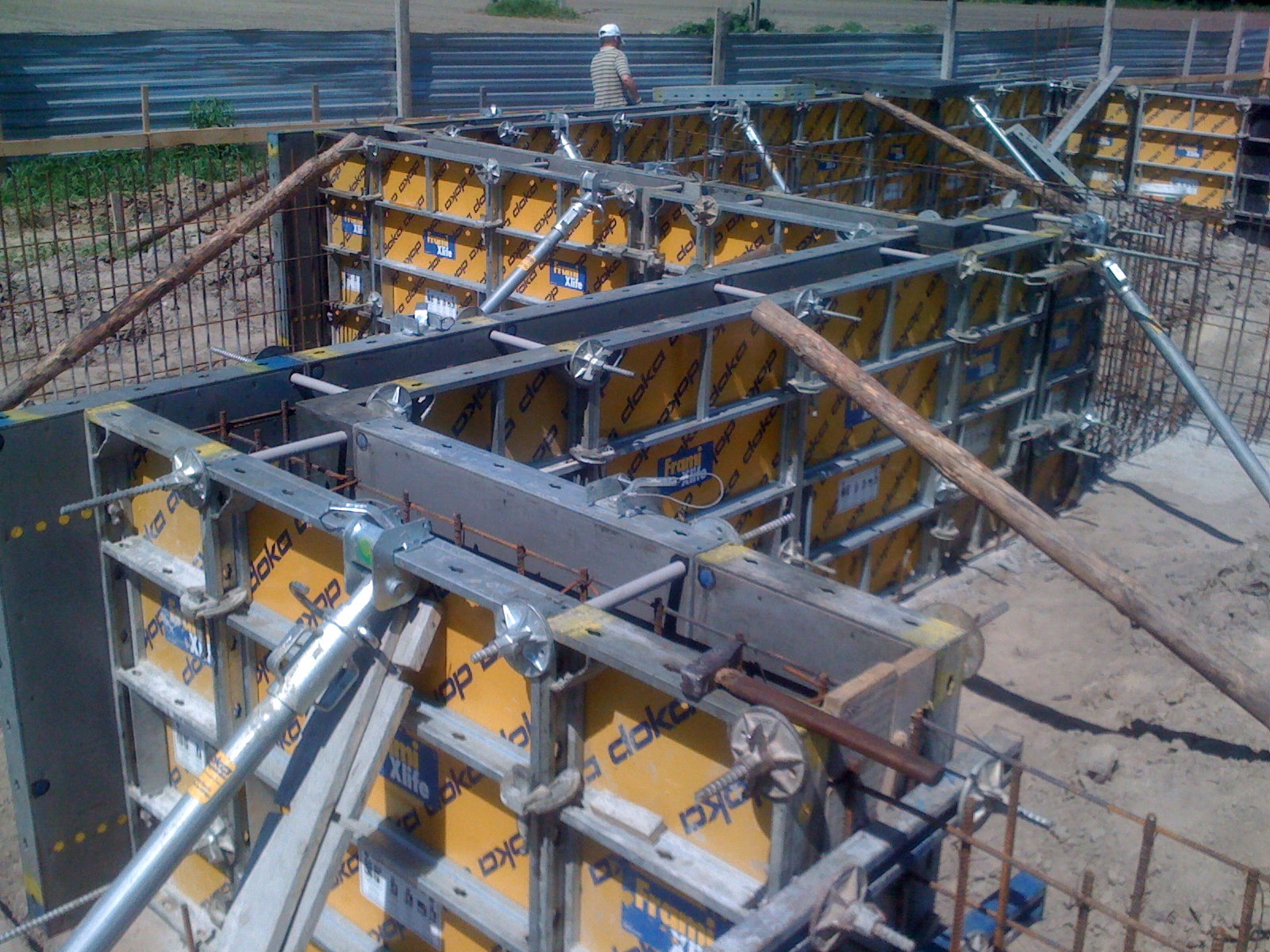 Szare szalunki stalowe z żółtymi panelami Doka Xlife, wzmocnione drewnianymi stemplami, przygotowane do wylania fundamentów; w tle widoczna siatka zbrojeniowa i pracownik w kasku.