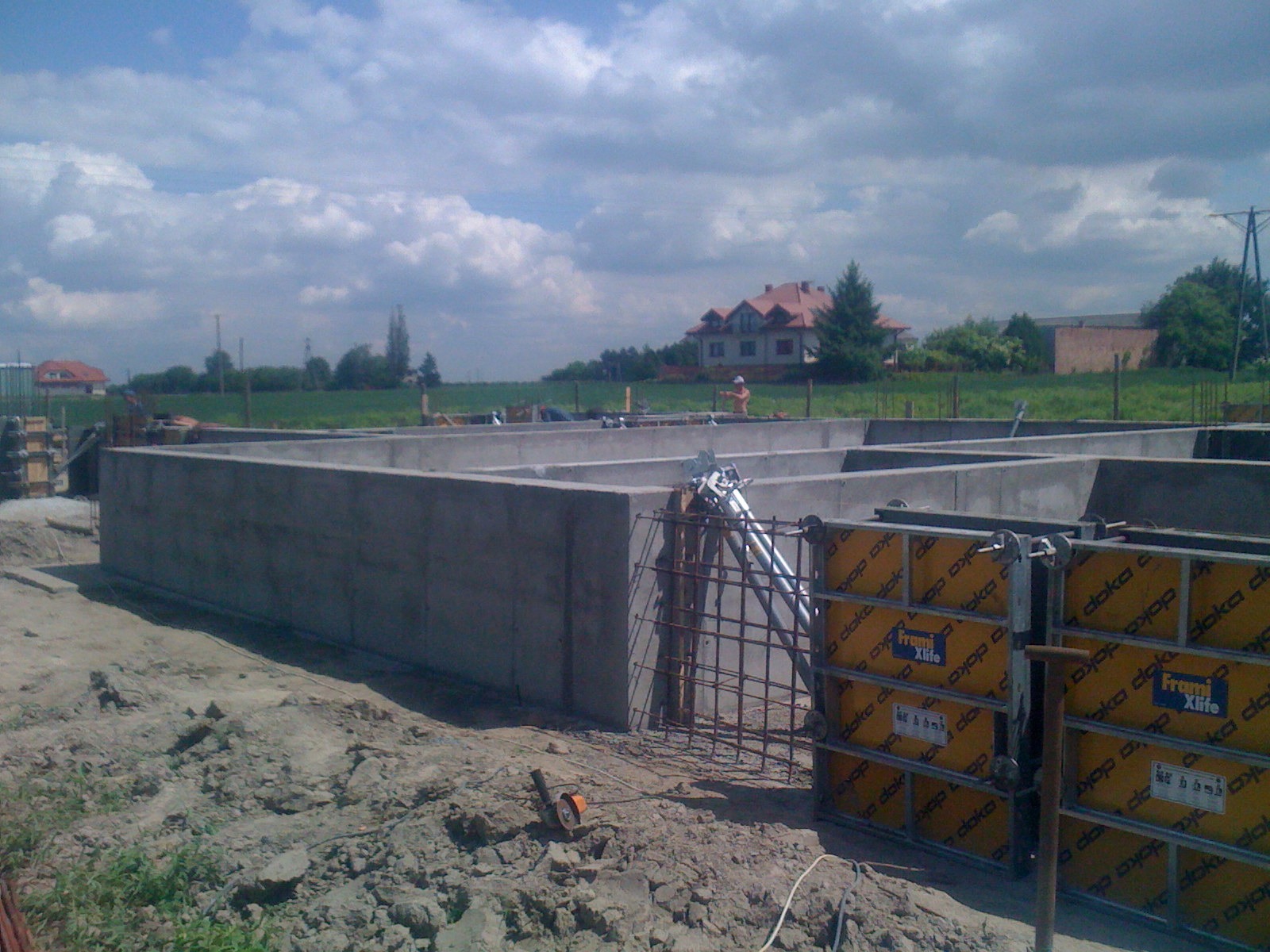 Wylane betonowe fundamenty domu jednorodzinnego, widoczne szalunki i zbrojenia, w tle budynek w trakcie budowy i pochmurne niebo.