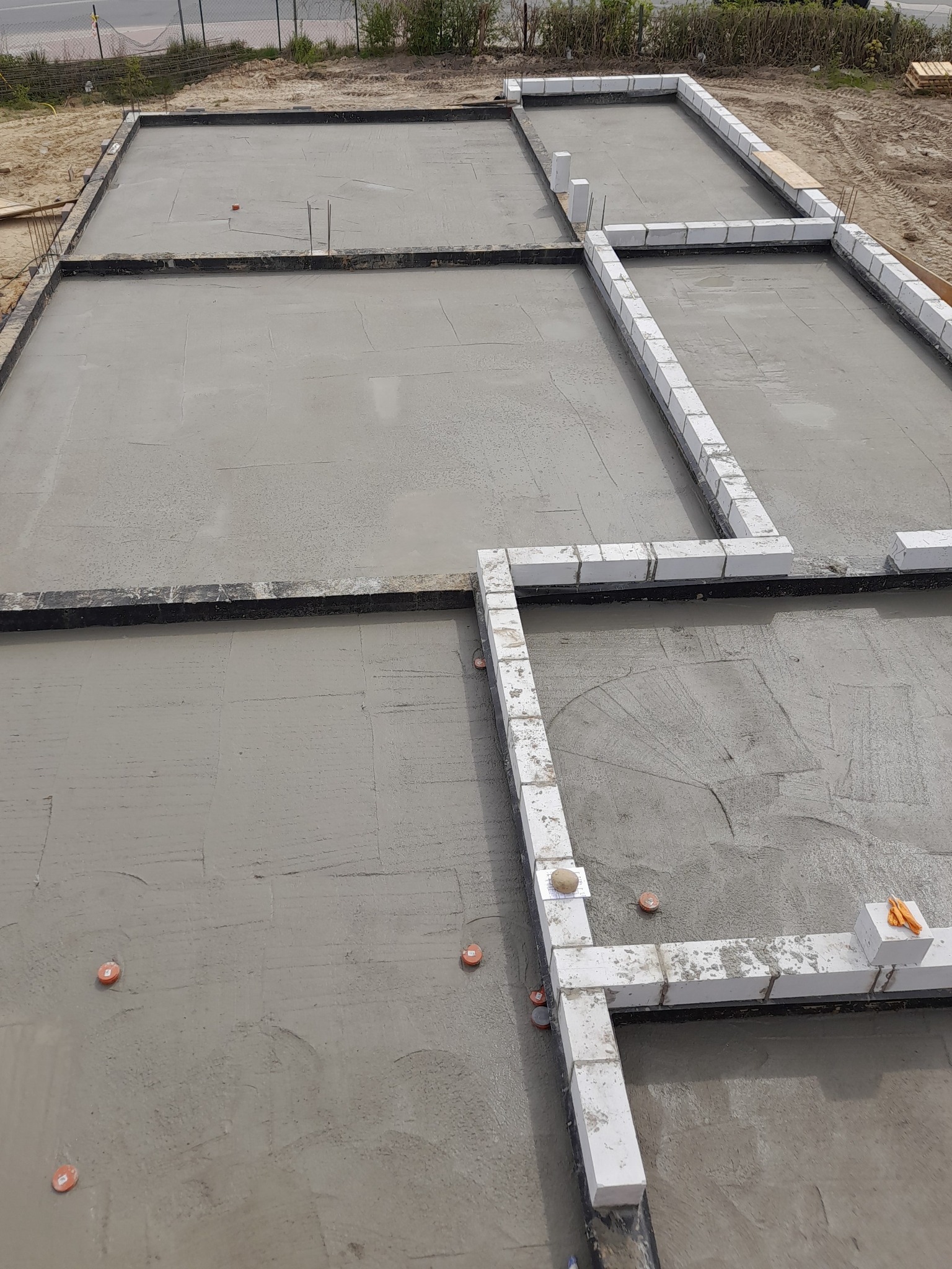 Zalany betonowy fundament z wyznaczonymi strefami pod ściany z bloczków Ytong, widoczne pomarańczowe znaczniki i wystające pręty zbrojeniowe.