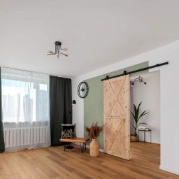 Good home - Jasne wnętrze z drewnianą przesuwną drzwią stodołową na czarnym szynoprzewodzie, zieloną ścianą akcentującą, drewnianą podłogą, ciemnozielonymi zasłonami i nowoczesnym oświetleniem.