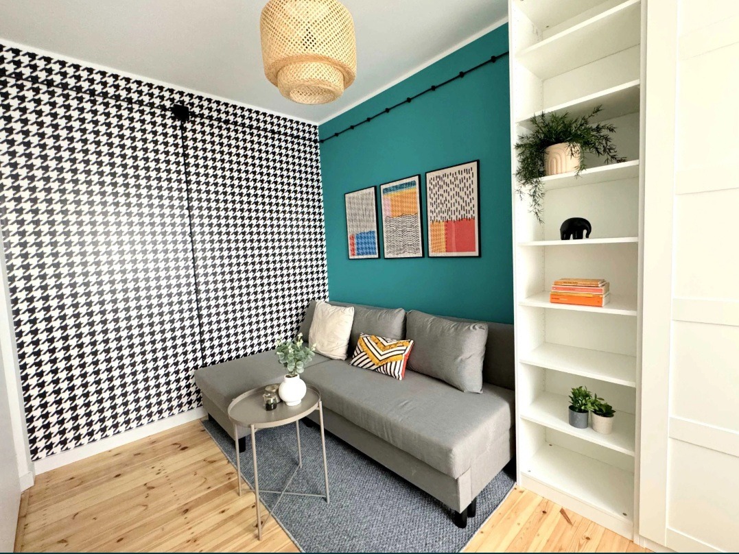 Nowoczesny pokój dzienny z szarą sofą narożną, stolikiem kawowym, wzorzystą tapetą w pepitkę, turkusową ścianą z trzema obrazami, rattanową lampą i białym regałem z dekoracjami.