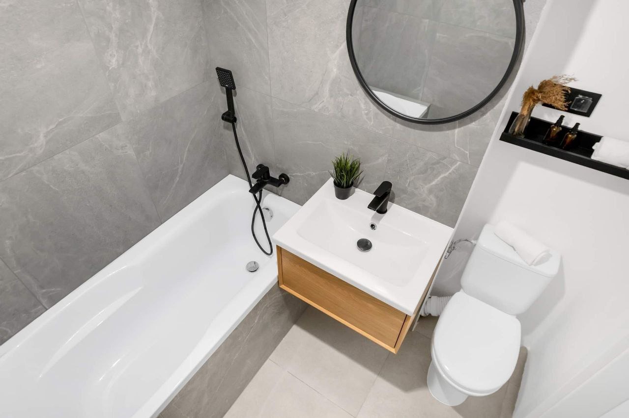 Nowoczesna łazienka z szarymi marmurowymi płytkami, białą wanną, wiszącą szafką z umywalką z drewnianym frontem, czarną armaturą i okrągłym lustrem.