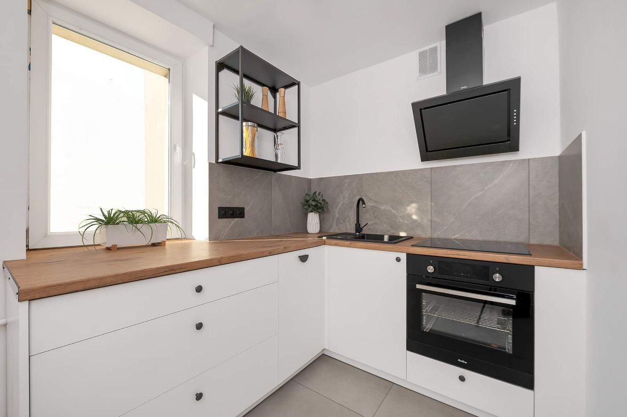Nowoczesna, jasna kuchnia z białymi szafkami, drewnianym blatem i szarymi płytkami imitującymi marmur. Na blacie roślina w doniczce, na ścianie czarna, metalowa półka z dekoracjami i czarny okap...