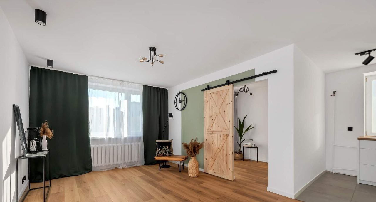 Jasne wnętrze z drewnianą przesuwną drzwią stodołową na czarnym szynoprzewodzie, zieloną ścianą akcentującą, drewnianą podłogą, ciemnozielonymi zasłonami i nowoczesnym oświetleniem.