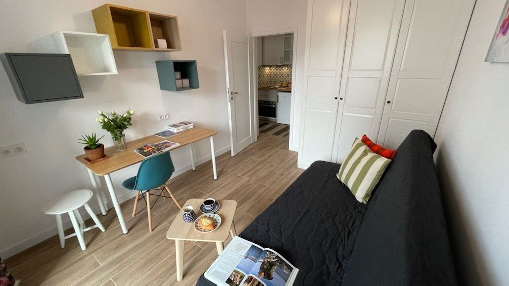 Jasne wnętrze pokoju po remoncie z drewnianą podłogą, biurkiem z kwiatami, kanapą z poduszkami, szafą wnękową i otwartą kuchnią w tle.