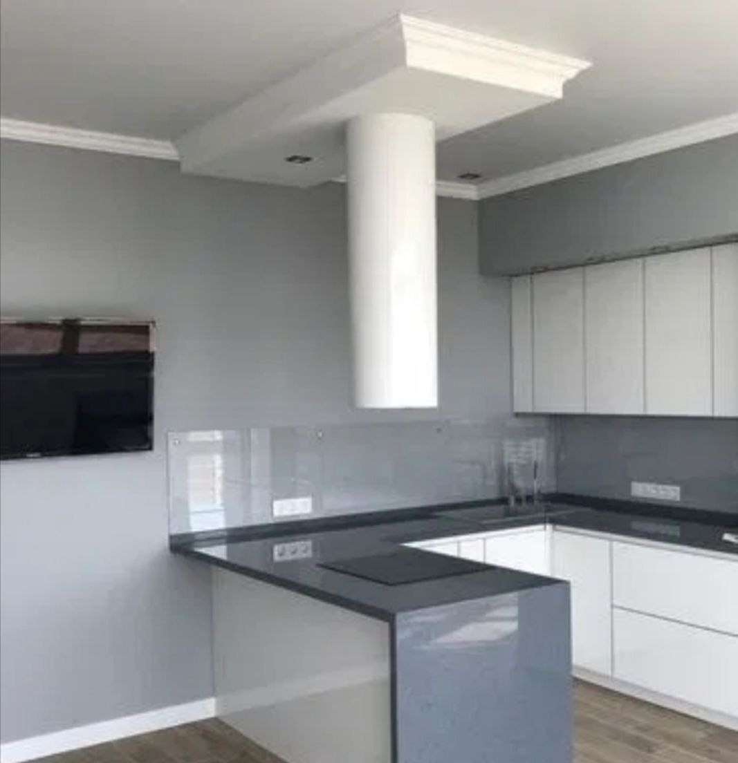 Nowoczesna kuchnia w odcieniach szarości i bieli z nietypowym okapem w kształcie cylindra, zamontowanym centralnie nad płytą grzewczą; widoczna szklana ściana nad blatem i telewizor na ścianie.