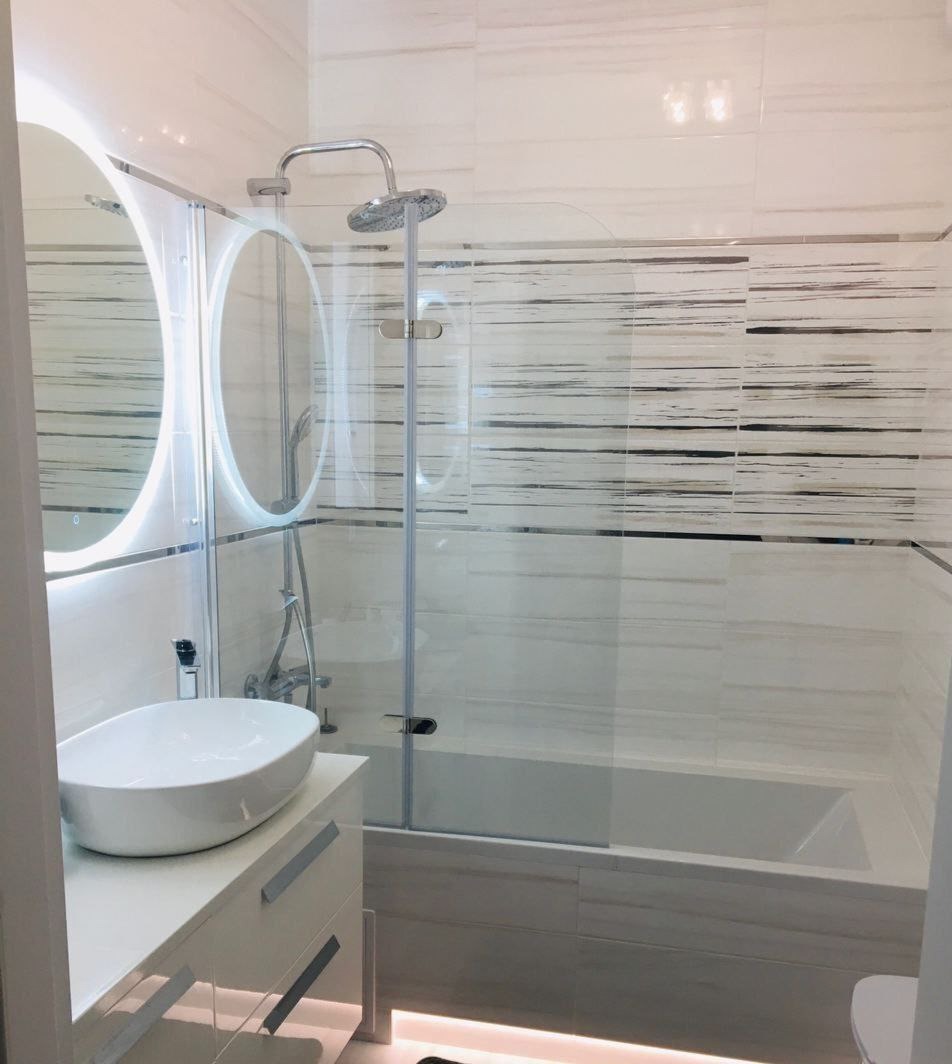 Nowoczesna łazienka z owalną umywalką na białej szafce z podświetleniem LED, kabina prysznicowa z szklanym panelem nad wanną i okrągłe lustra z podświetleniem LED na tle płytek w poziome pasy.