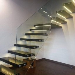 Schody System - Nowoczesne schody z drewnianymi stopniami podświetlonymi LED i szklaną balustradą, wsporniki mocujące stopnie do ściany.