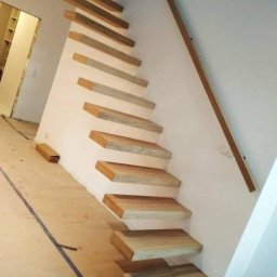 Schody System - Nowoczesne, minimalistyczne schody drewniane wspornikowe z drewnianą poręczą zamontowane przy białej ścianie, widoczne ślady prac wykończeniowych na podłodze.